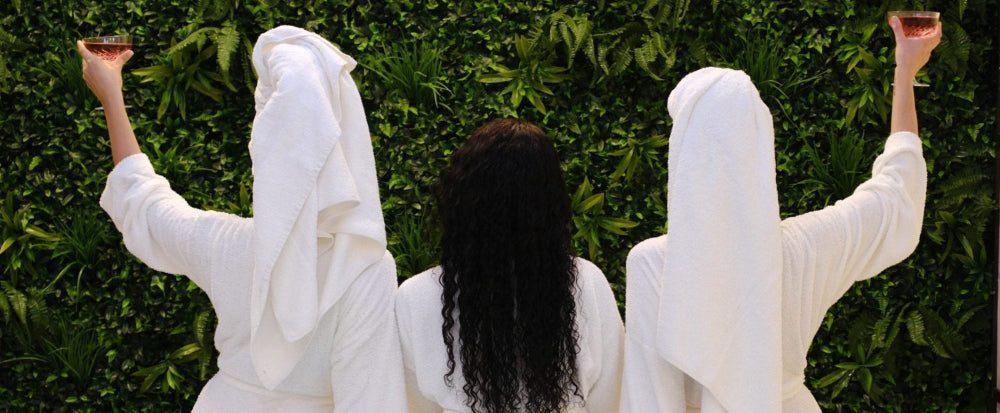 Three people wrapped in white towels by a pool, holding drinks.